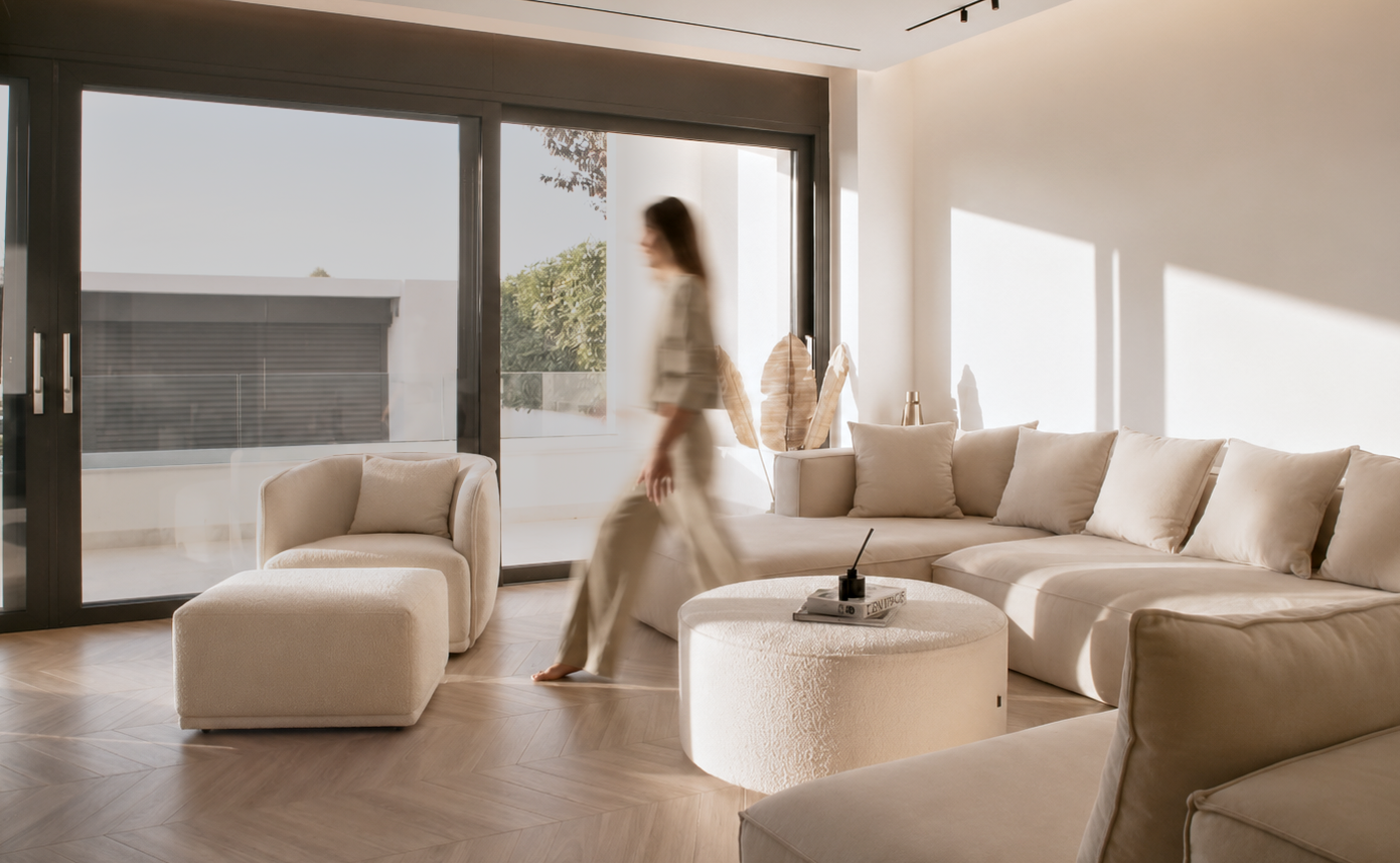 Sunlit contemporary living room in Thessaloniki with a figure walking past floor-to-ceiling glazing